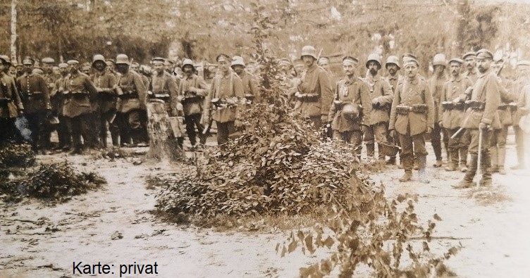 Das Foto zeigt eine Gruppe von Soldaten, die in Uniformen aus der Zeit des Ersten Weltkriegs posieren. Sie tragen Stahlhelme und halten Gewehre bei sich. Die Männer stehen im Freien in einer Waldlandschaft. Im Vordergrund ist ein Haufen Laub oder möglicherweise eine Tarnung zu sehen. Unten auf dem Foto steht "Karte: privat".