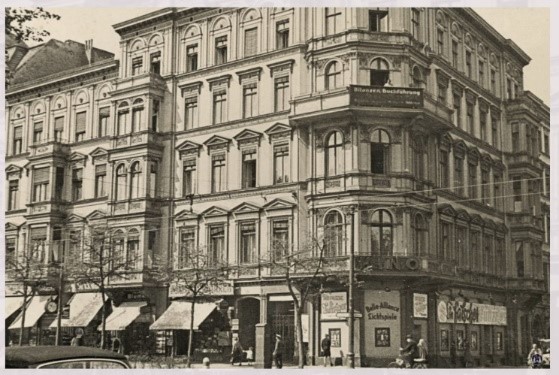 Das Foto zeigt ein eckiges Gebäude im Stil der Gründerzeit mit mehreren Stockwerken und großen Fenstern. Im Erdgeschoss sind Geschäfte mit Markisen und Werbeschildern. An der Ecke des Gebäudes befindet sich ein runder Balkon. Die Architektur weist auf ein städtisches Umfeld hin, vermutlich in Deutschland aus der ersten Hälfte des 20. Jahrhunderts.