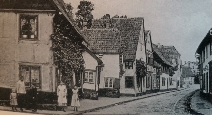 Das Bild zeigt eine alte Straße mit Kopfsteinpflaster und traditionellen, giebelständigen Häusern. Links sind fünf Kinder zu sehen.