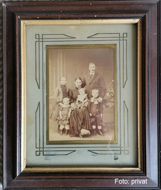 Das Foto zeigt ein viktorianisches Familienporträt, gerahmt in einem dunklen Bilderrahmen mit geometrischen Art-Deco-Verzierungen. Eine Familie von sechs Personen, zwei Erwachsene und vier Kinder, posieren gemeinsam. Der Mann und die Frau sitzen in der Mitte, er in dunklem Anzug und Schnurrbart, sie in einem dunklen Kleid mit weißer Bluse, ein Buch haltend. Die Kinder stehen um sie herum, zwei Jungen in dunklen Anzügen und ein Mädchen in einem hellen Kleid. Unten steht "Foto: privat".