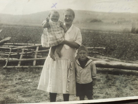 Eine Frau steht in der Mitte des Bildes. Sie hält ein kleines Mädchen auf dem Ar,m. Rechts von ihr steht ein Junge in einem Matrosenanzug. Hinter ihnen ein Acker, davor liegt Holz auf dem Boden